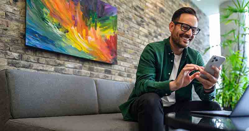 Man in a green shirt looking at his phone while sitting on a couch with a colorful oil painting on the wall behind him.